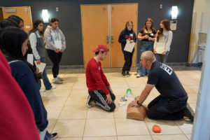 Several students st6and and watch a Protection 1 EMS employee demonstrate CPR on a dummy.