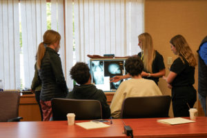 Several students stand around an x-ray while a presenter4 points to a part of the x-ray to explain to the students.