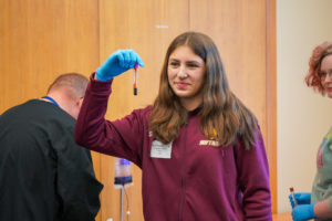 A girl in a purple sweater holds up a vial of blood.