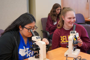 One student looks into a microscope while a second student smiles.