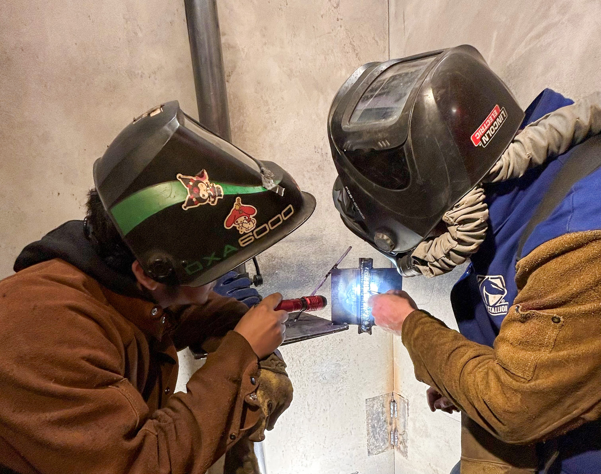 A student on the left holds a flashlight while an instructor on the right points at a part of the student's weld work.
